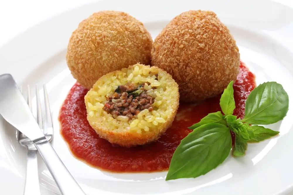 Zlatavé smažené sicilské arancini s rýžovou náplní a mletým masem, podávané s rajčatovou omáčkou a čerstvou bazalkou na bílém talíři