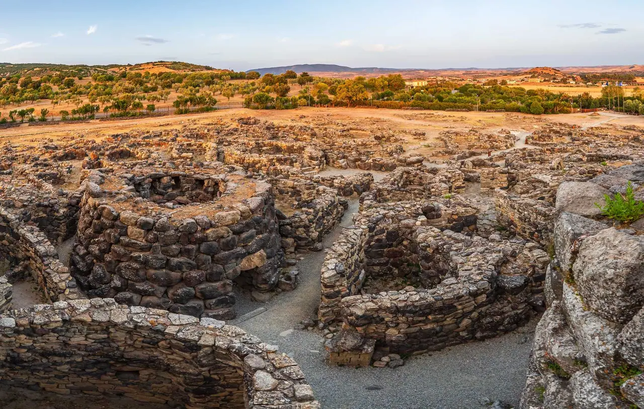 Pohled shora na kamenné věžové stavby nuraghe Su Nuraxi v Barumini na Sardinii, obklopené suchými pahorky s olivovníky.
