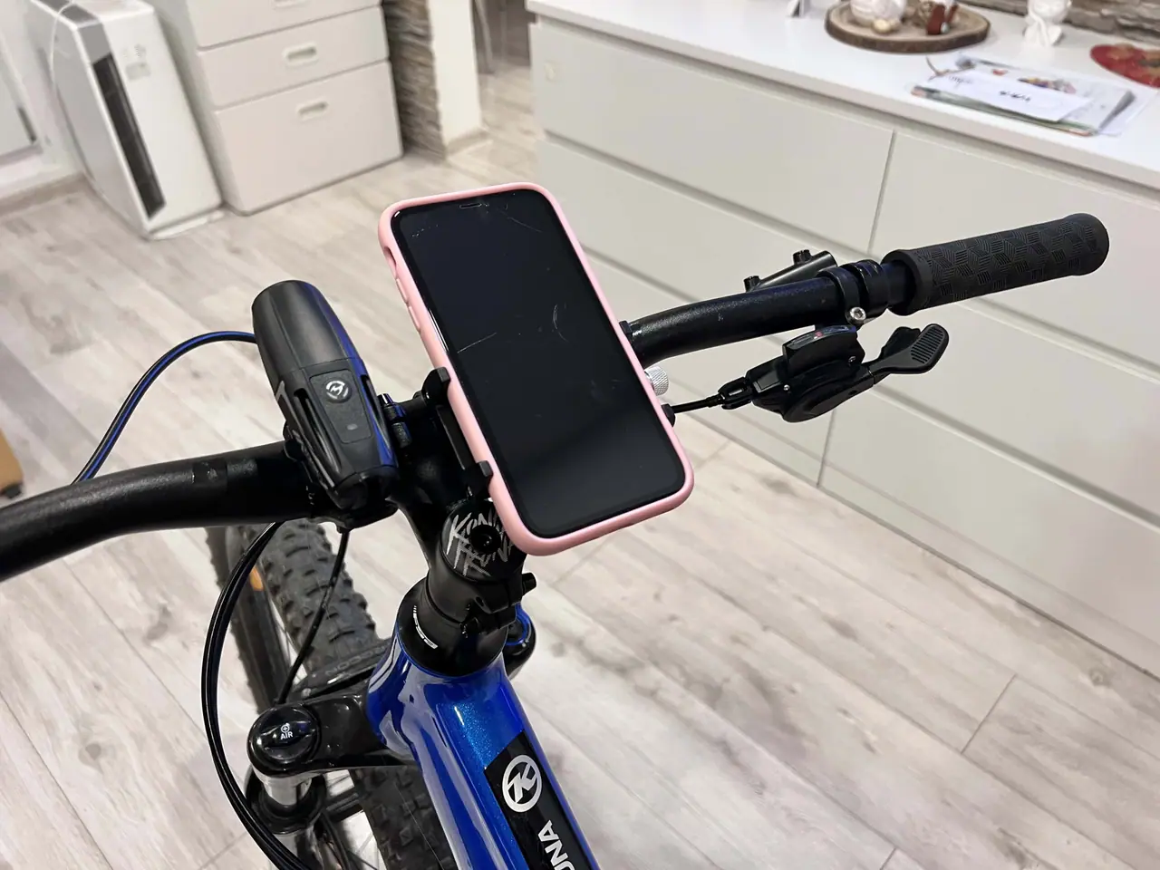 Kovový držák telefonu Swissten BC2 namontovaný na řídítkách modrého kola s růžovým mobilem v kuchyni