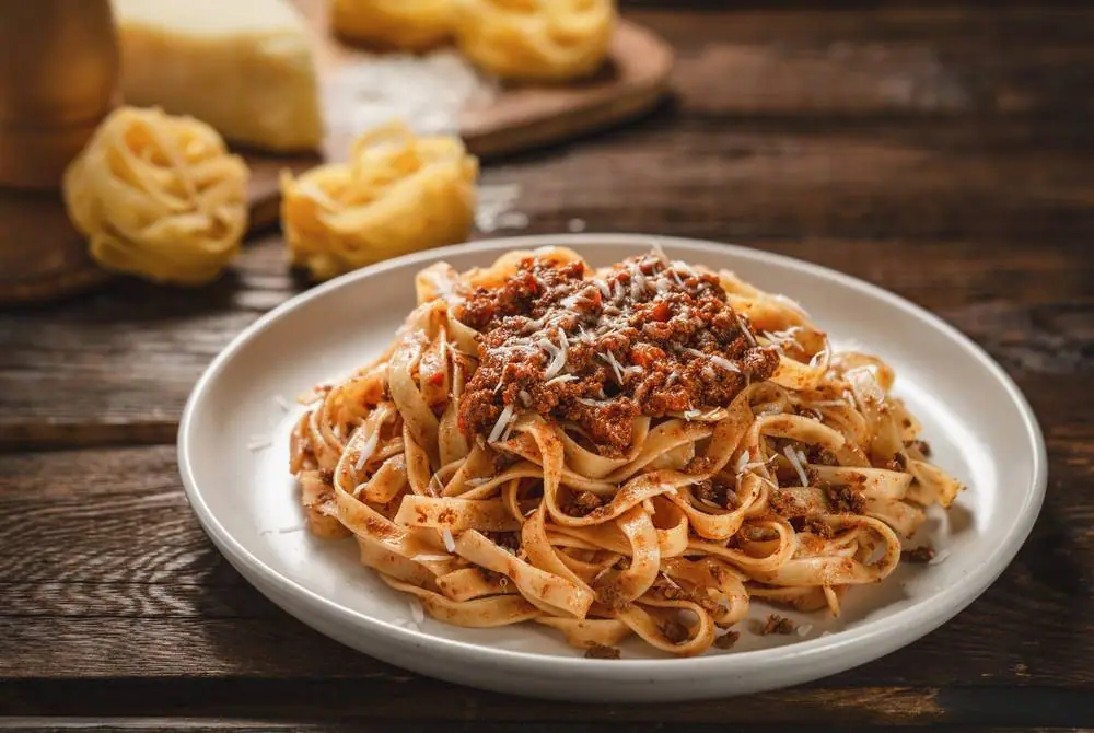 Čerstvě uvařené tagliatelle s boloňskou omáčkou na bílém talíři, kolem rozložené sušené nudle a parmazán na dřevěném stole