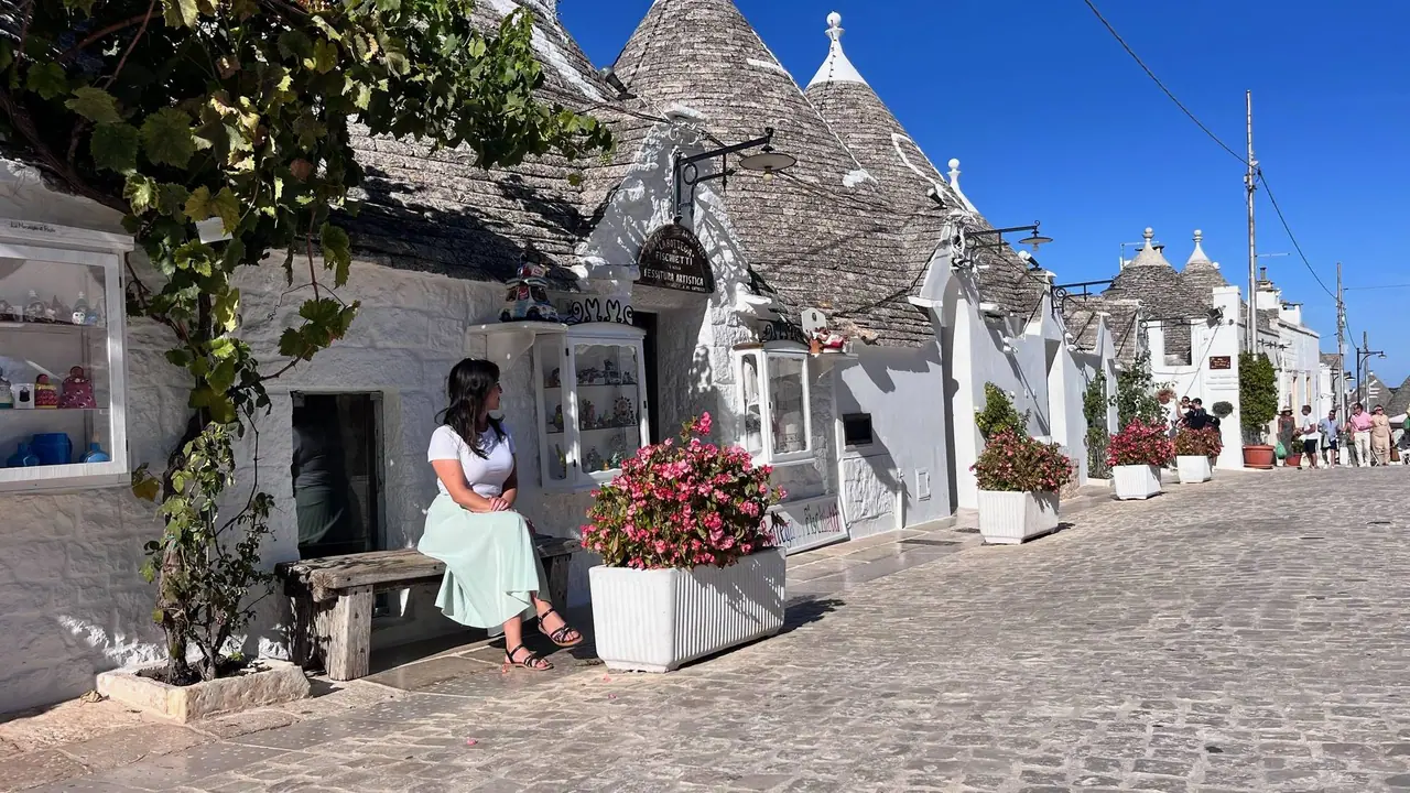 Žena v bílém tričku a světle zelené sukni sedí na lavičce před tradičními trulli domky s kuželovitými kamennými střechami v Alberobello