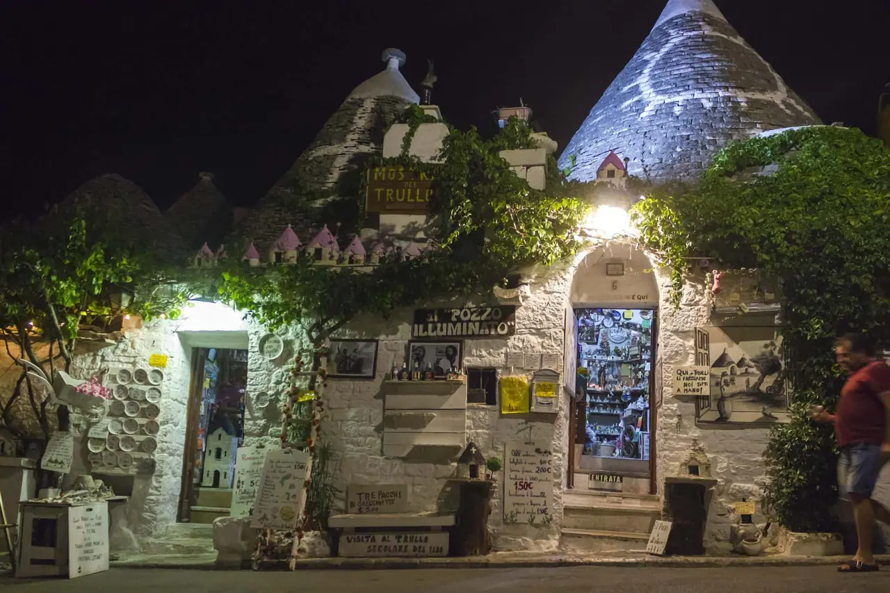 Noční pohled na osvětlené trulli domky v Alberobello s kamenými kužely, suvenýrovými obchůdky a turistickými ceduleми pod tmavou oblohou