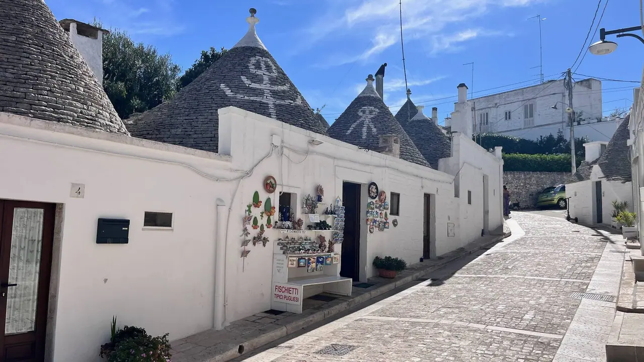 Bílé trulli s kamennými kuželovitými střechami v úzké dlážděné uličce Alberobella, na jednom domku visí suvenýry a pohlednice