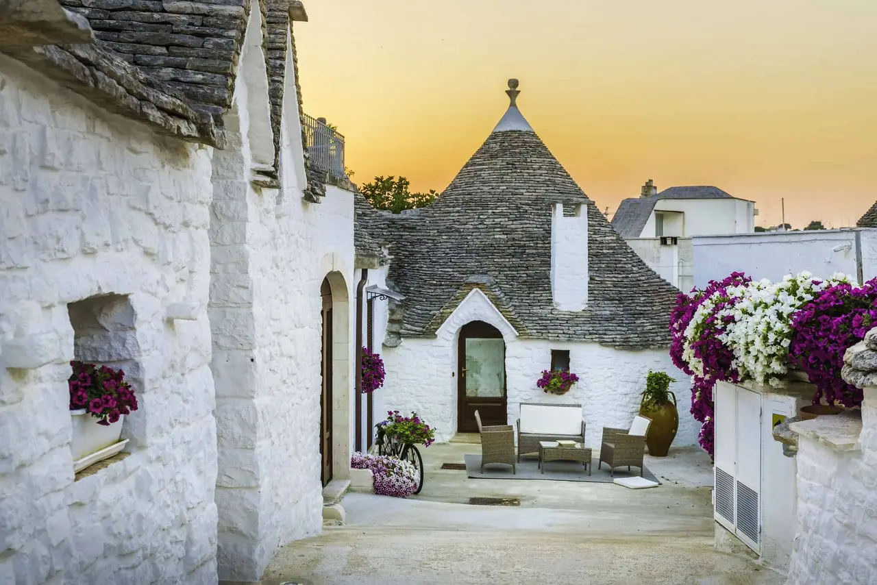 Bílé trulli s kuželovými střechami z kamene v Alberobello za soumraku, fialové květiny na terasách a moderní zahradní nábytek