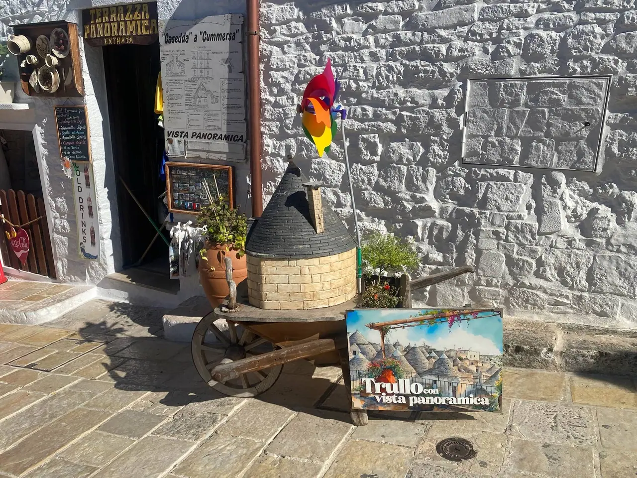 Dřevěný vozík s modelem trullo a suvenýry před kamennou zdí v Alberobello, barevná vlajka vlaje ve větru