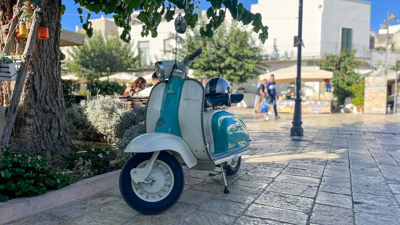 Vintage tyrkysová Vespa Lambretta zaparkovaná na náměstí v historickém centru Alberobella s trulli domečky v pozadí