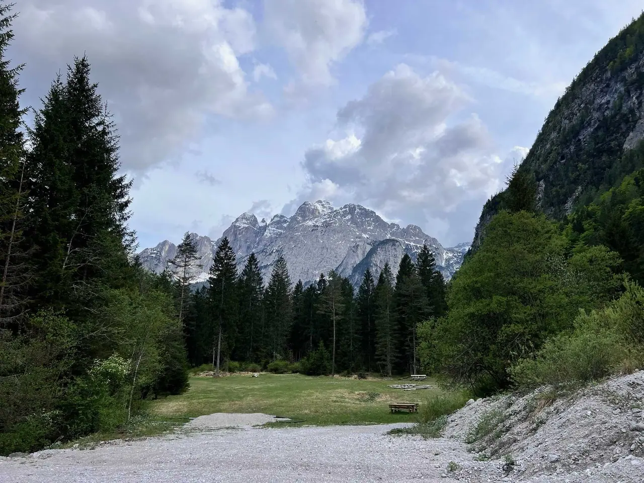 Štěrkové parkoviště u údolí Val Saisera s dřevěnými lavičkami na travnaté louce, obklopené jehličnatými stromy a skalnatými vrcholky Julských Alp s posledními zbytky sněhu