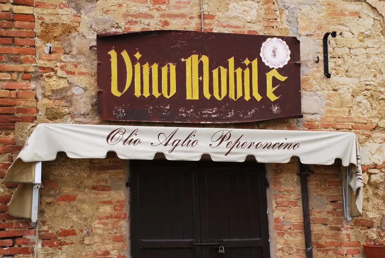 Vstup do prodejny Vino Nobile v Montepulciano s tmavě červenou cedulí a bílou markýzou nad dřevěnými dveřmi ve zdi z cihel a kamene