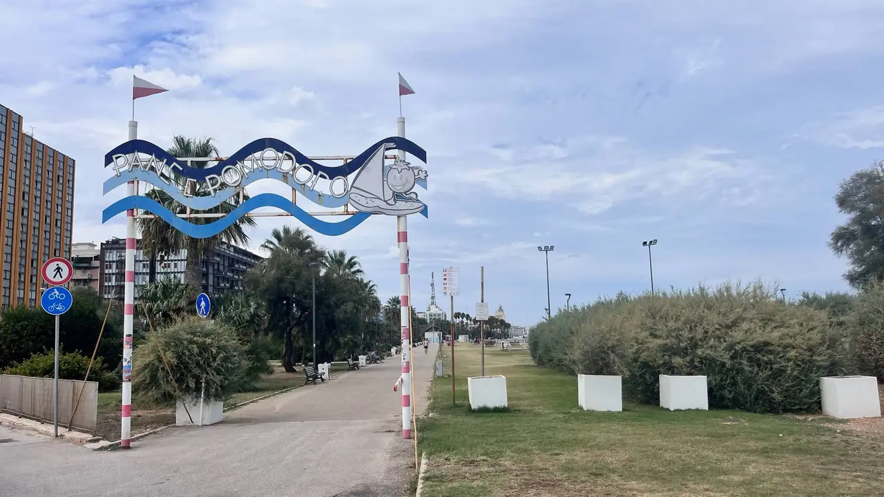 Vstupní brána do Bari s nápisem a vlajkami, vedoucí k pobřežní promenádě s palmami a hotely v pozadí